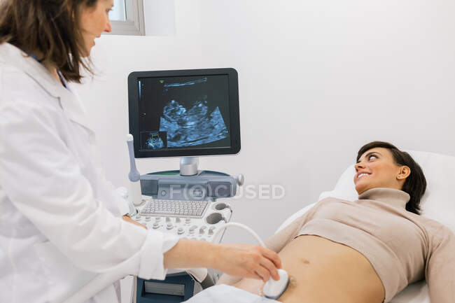 Médico de cultura fazendo sonograma para a mulher grávida feliz durante o trabalho na clínica de fertilidade contemporânea — Fotografia de Stock