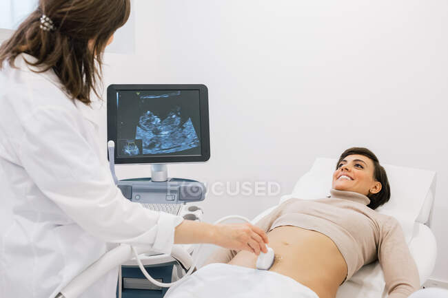 Médico de cultura fazendo sonograma para a mulher grávida feliz durante o trabalho na clínica de fertilidade contemporânea — Fotografia de Stock
