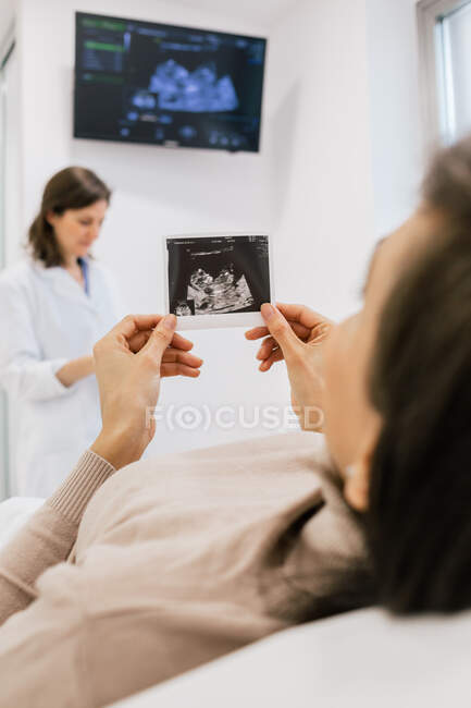 Mulher grávida irreconhecível inspecionando imagem de ultrassonografia enquanto deitada na cama na enfermaria da clínica de fertilidade moderna — Fotografia de Stock