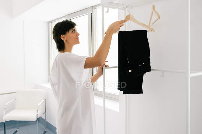 Vista lateral de la mujer feliz en bata blanca colgando pantalones en el carril mientras se desnuda antes del procedimiento médico en la sala de la clínica - foto de stock
