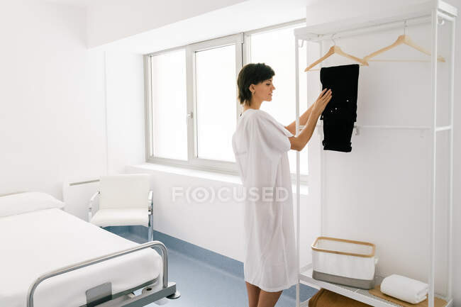 Side view of happy female in white robe hanging pants on rail while undressing before medical procedure in clinic ward — Stock Photo