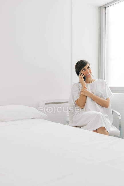 Mulher alegre em roupão branco sentado na cama perto da cama e usando smartphone na ala da clínica moderna — Fotografia de Stock