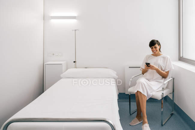 Cheerful woman in white robe sitting on bed near bed and browsing smartphone in ward of modern clinic — Stock Photo