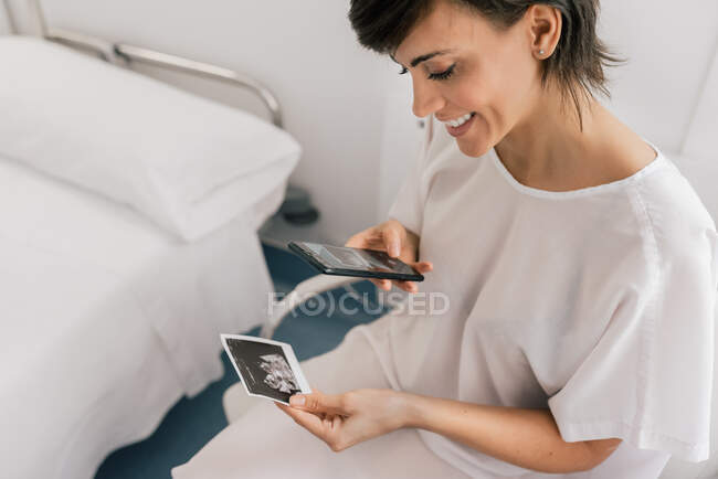 From above cheerful female patient smiling and taking picture of ultrasound scan while sitting in ward of fertility clinic — Stock Photo