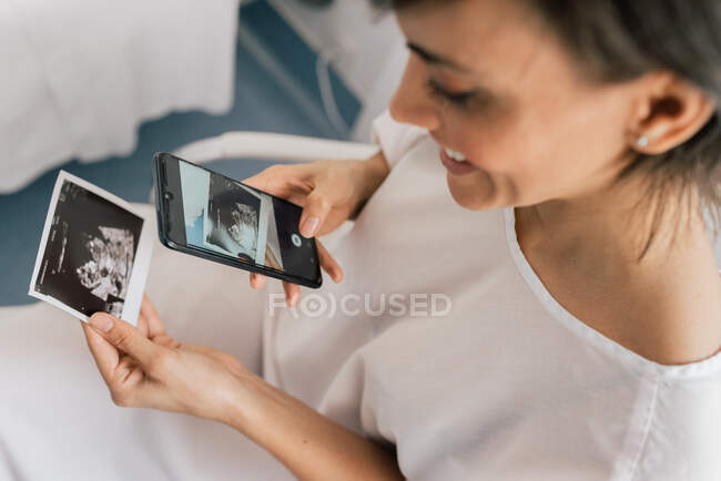 D'en haut joyeuse patiente souriante et prenant des photos de l'échographie tout en étant assise dans la salle de la clinique de fertilité — Photo de stock