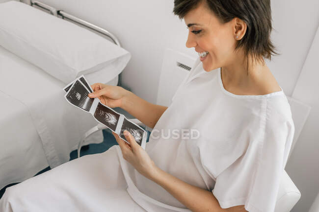 De arriba embarazada hembra inspeccionando imagen de ecografía mientras está sentado en una silla en la sala de la clínica de fertilidad moderna - foto de stock