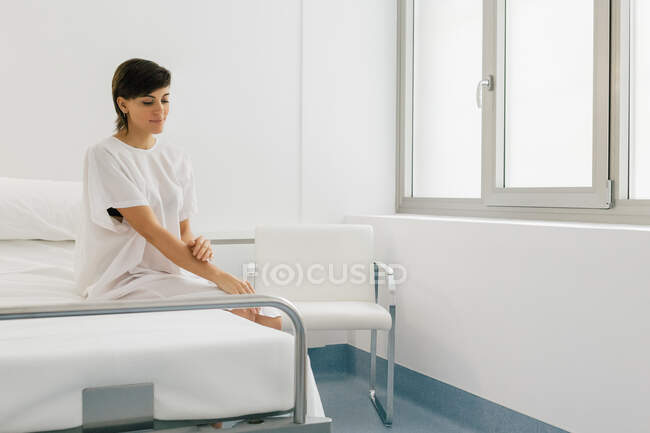 Радостная женщина в белом халате улыбается и сидит на кровати в светлом отделении современной клиники — стоковое фото