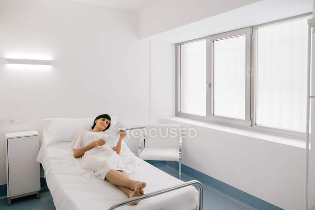 Вагітна жінка оглядає фотографію сонограми, лежачи на ліжку в палаті сучасної клініки фертильності — стокове фото