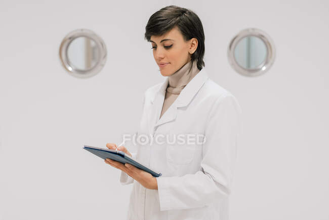 Side view of positive medical practitioner using tablet while standing against white door in hallway of modern hospital — Stock Photo