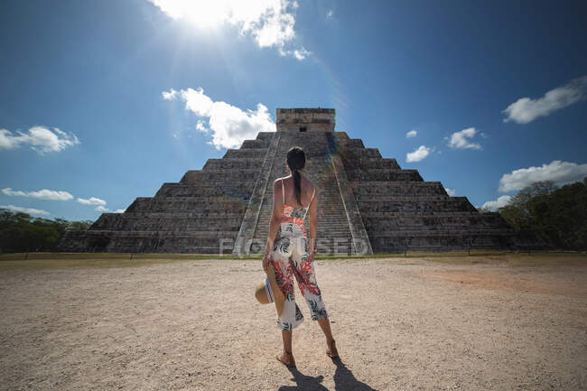 Visão traseira do viajante feminino anônimo em roupas brilhantes de verão segurando chapéu na mão e admirando a vista do antigo templo de pedra durante as férias no ensolarado México — Fotografia de Stock