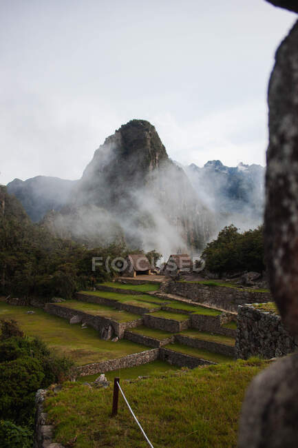 Удивительный вид на зеленую скалистую долину с каменными заборами и маленькими каменными домами, расположенными вблизи пика, покрытого туманными облаками в Перу — стоковое фото