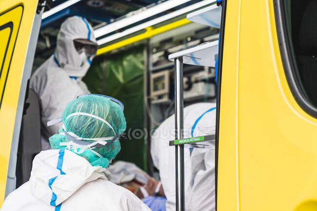 Vista lateral de los médicos anónimos en traje de protección de pie en el coche de ambulancia cerca de la puerta abierta con el equipo y examinar al paciente con infección por virus - foto de stock