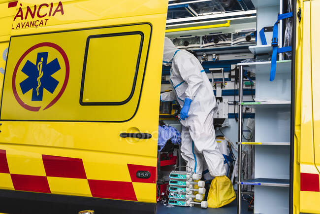 Vista lateral del médico anónimo en traje de protección de pie en el coche de ambulancia cerca de la puerta abierta con el equipo y examinar al paciente con infección por virus - foto de stock