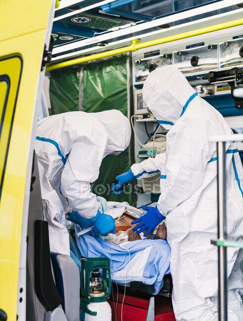 Vista lateral de médicos anônimos em terno de proteção em pé no carro de ambulância perto da porta aberta com equipamento e examinando o paciente com infecção pelo vírus — Fotografia de Stock