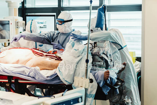 Médicos profissionais irreconhecíveis em uniformes e máscaras protetoras cuidando de pacientes com infecção viral em pé na sala de cirurgia no hospital moderno — Fotografia de Stock