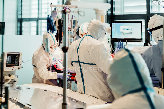 Неузнаваемые профессиональные врачи в защитной форме и масках, ухаживающие за пациентом с вирусной инфекцией, стоя в операционной в современной больнице — стоковое фото