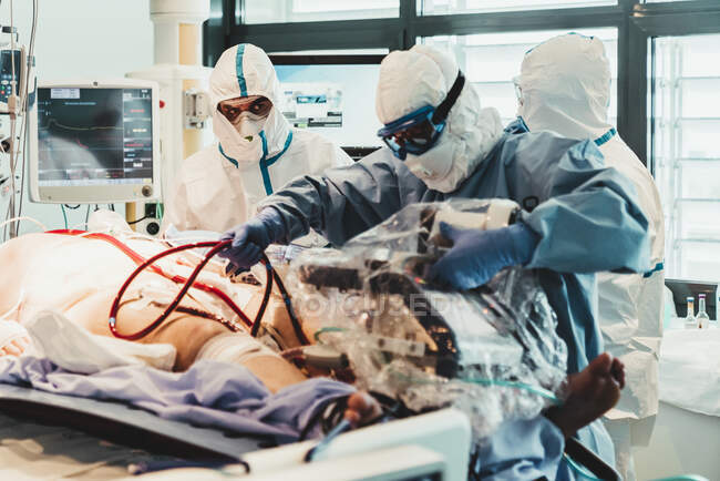 Médicos profesionales irreconocibles con uniformes protectores y máscaras que cuidan al paciente con infección viral mientras está de pie en el quirófano en el hospital moderno - foto de stock