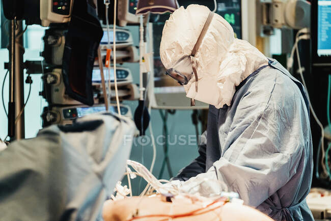 Vista lateral del médico profesional irreconocible en uniformes protectores y máscaras cuidando al paciente con infección viral mientras está de pie en el quirófano en el hospital moderno - foto de stock