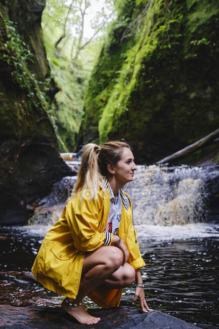 Vista lateral de la mujer viajera feliz en chaqueta amarilla agachándose y tocando piedra cerca de la cascada que pasa entre las montañas a la luz del sol mirando hacia otro lado - foto de stock