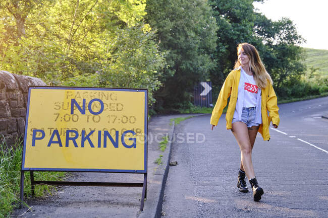Счастливая женщина в желтой куртке и джинсовых шортах, смотрящая на дорожный знак — стоковое фото