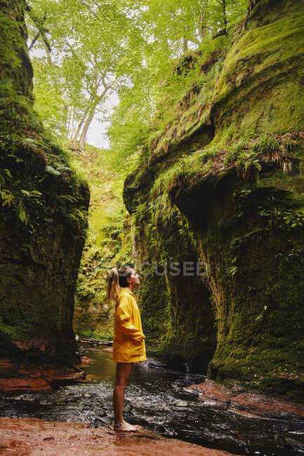 Vue latérale du voyageur féminin en veste jaune debout dans la pierre près de la cascade passant entre les montagnes en plein soleil regardant loin en admirant la vue — Photo de stock