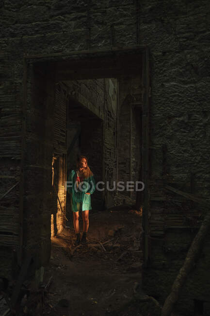 Femme blonde en manteau vert et bottes marchant les mains dans la poche dans un ancien bâtiment sombre au coucher du soleil — Photo de stock