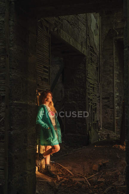 Side view of blond female in green coat and boots leaning on wall with hands in pocket while standing near dark ancient building at sunset — Stock Photo