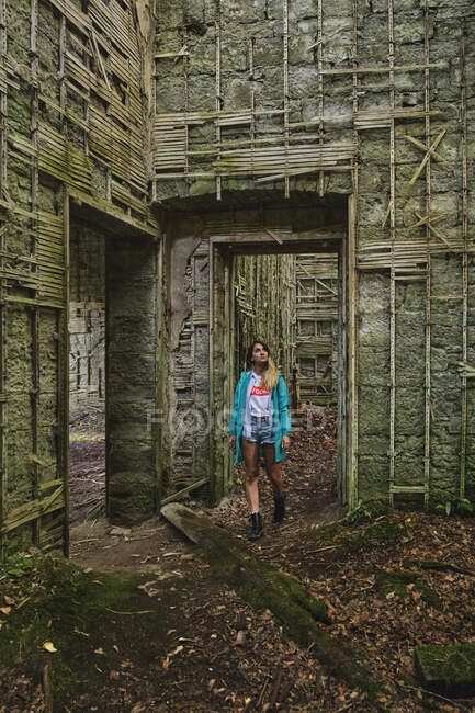 Viaggiatore donna in cappotto blu e stivali che cammina su un sentiero coperto di foglie appassite mentre osserva antiche costruzioni fatte di pietre e bastoni di legno. — Foto stock