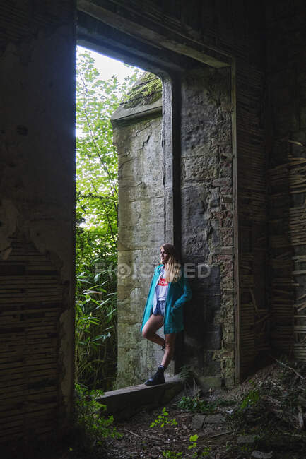 Счастливая женщина-путешественница в синем пальто и сапогах, опирающаяся на стену древней конструкции из камней и деревянных палок, отворачивающихся — стоковое фото