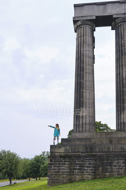 Side view of woman in colorful jacket standing on memorial monument steps and pointing with finger at sky on sunny day in park — Stock Photo