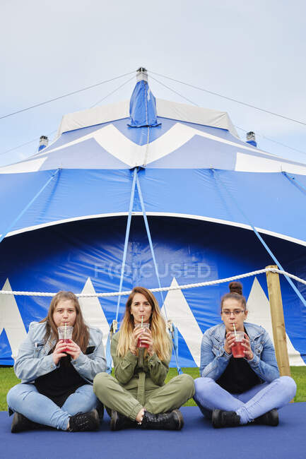 Cheerful young women in casual clothes drinking red beverage through straw while sitting on blue mat near fenced tent — Stock Photo