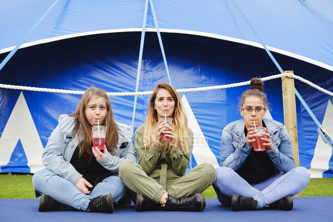 Jovens mulheres alegres em roupas casuais bebendo bebida vermelha através de palha enquanto sentado no tapete azul perto da tenda cercada — Fotografia de Stock