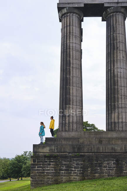 Vista lateral das mulheres em casacos coloridos em pé nos degraus do monumento memorial e apontando com o dedo para o céu no dia ensolarado no parque — Fotografia de Stock