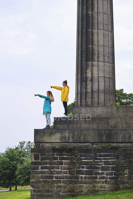 Side view of women in colorful jackets standing on memorial monument steps and pointing with finger at sky on sunny day in park — Stock Photo