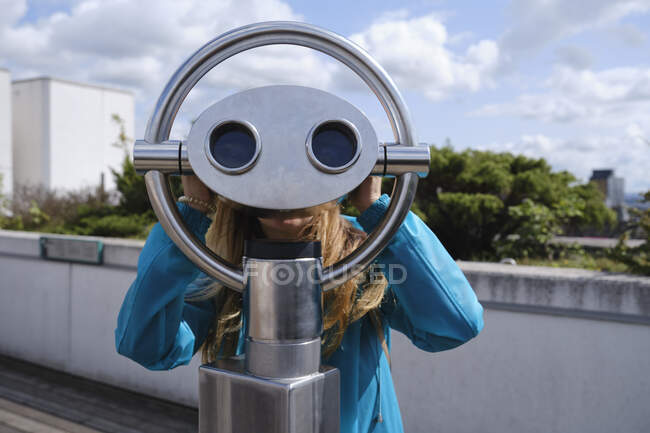 Anonymous blond female in blue coat looking through street coin operated binocular while standing on wooden pavement near concrete fence on sunny day — Stock Photo