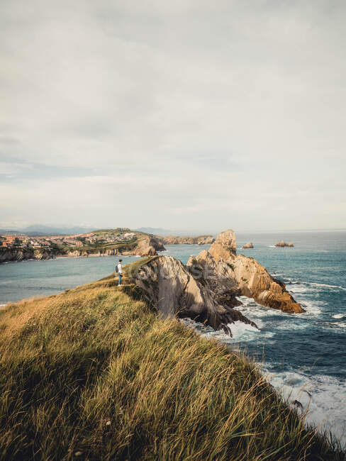 Vista lateral distante do viajante masculino com mochila e câmera em pé na costa rochosa e admirando paisagens marinhas incríveis durante a viagem na Espanha — Fotografia de Stock