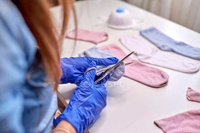 Dall'alto di femmina anonima con i capelli rossi in blu guanti sterili taglio calzino con le forbici a casa durante la fabbricazione di maschera durante COVID 19 — Foto stock