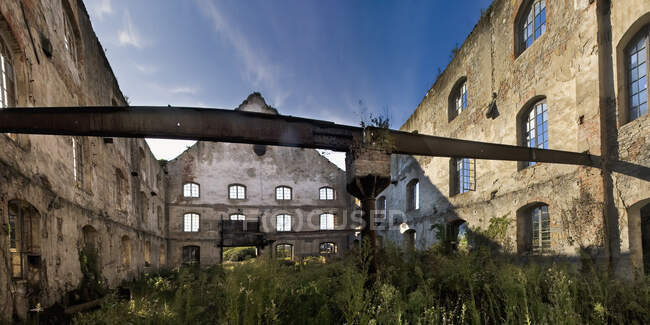 Bajo ángulo de paredes de piedra envejecida de antiguo edificio industrial con ventanas enrejadas y suelo cubierto contra cielo azul en día soleado en Asturias en España - foto de stock