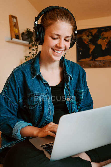Mujer pensativa alegre en auriculares y ropa casual que se centra en la pantalla con interés y el uso de netbook mientras está sentado con las piernas cruzadas en la cama suave en acogedor apartamento moderno - foto de stock