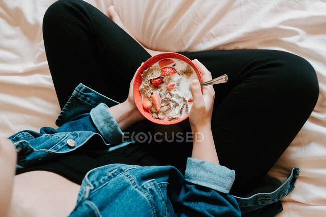 Von oben eine Frau in lässiger Kleidung mit einem Metalllöffel und einer roten Schüssel mit Erdbeerstücken und Haferflocken, die mit Milch übergossen werden, während sie im hellen Schlafzimmer auf dem Bett sitzt — Stockfoto