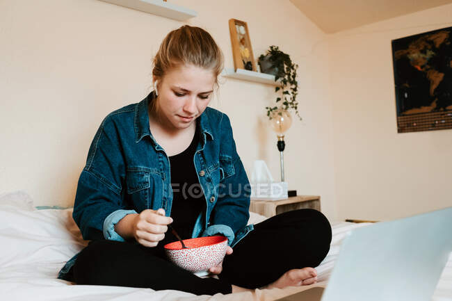 Ошеломленная задумчивая босиком женщина в настоящих беспроводных наушниках и повседневной одежде с миской вкусной еды, фокусирующейся на экране с интересом, сидя со скрещенными ногами на мягкой кровати и используя нетбук в уютной квартире — стоковое фото