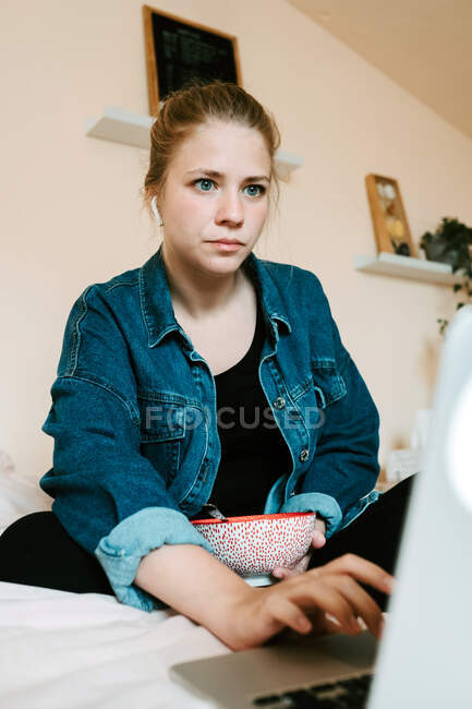 Stunned pensive barefoot female in true wireless earphones and casual clothes with bowl of tasty food focusing on screen with interest while sitting with legs crossed on soft bed and using netbook in cozy apartment — Stock Photo