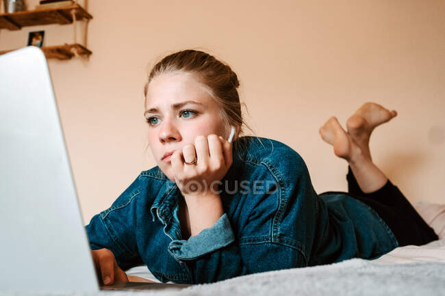 Thoughtful focused female in true wireless earbuds and casual clothes lying down on bed using laptop against beige wall in light apartment — Stock Photo