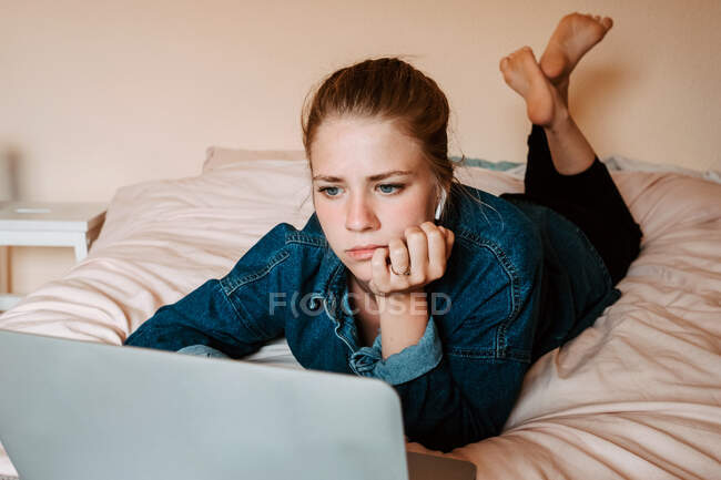 Nachdenkliche fokussierte Frau in echten drahtlosen Ohrhörern und lässiger Kleidung legt sich mit Laptop gegen beige Wand in heller Wohnung ins Bett — Stockfoto
