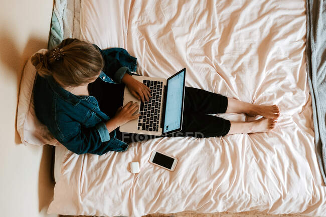 Зверху непізнавана босоніжка в повсякденному одязі, що фокусується на екрані та працює на ноутбуці, сидячи на м'якому ліжку зі смартфоном та бездротовими навушниками у затишній сучасній квартирі — стокове фото
