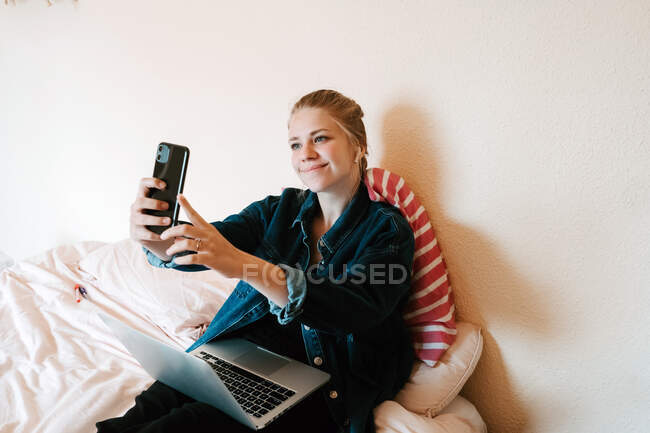 Молодая счастливая женщина в беспроводных наушниках и джинсовой куртке с помощью смартфона делает селфи и ноутбука, отдыхая на кровати в современной квартире — стоковое фото