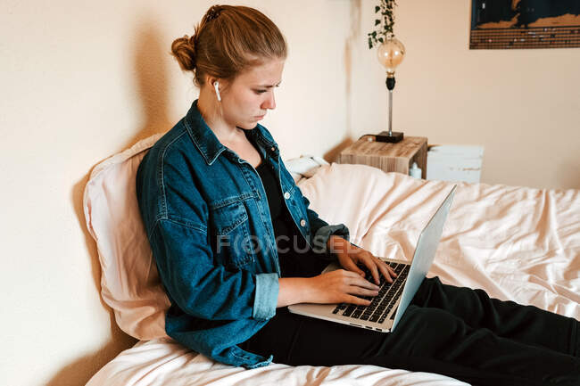 Side view of thoughtful focused female in true wireless earbuds and casual clothes sitting on bed using laptop against beige wall in light apartment — Stock Photo