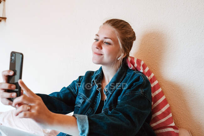 Радостная молодая женщина в беспроводных наушниках и джинсовой куртке улыбается на экране и делает селфи со смартфоном, отдыхая на кровати в современной квартире — стоковое фото