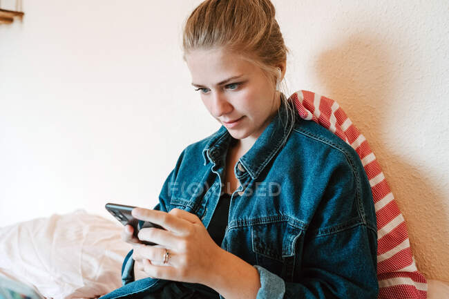 Молода жінка в бездротових навушниках і джинсовій куртці використовує смартфон, розслабляючись на ліжку в сучасній квартирі — стокове фото
