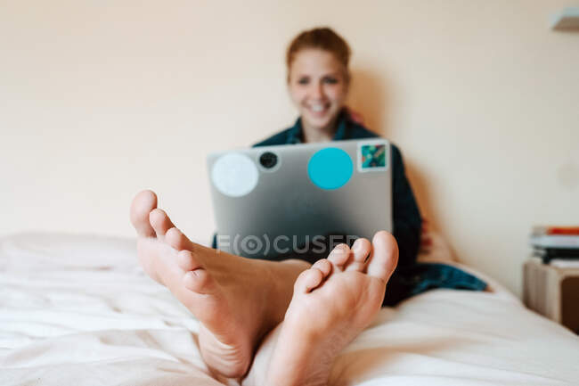 Blurred barefoot relaxed lady in casual clothes smiling at camera while sitting with legs crossed on soft bed and using netbook against light beige wall in cozy contemporary apartment — Stock Photo